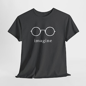 Puede incluir: Camiseta gris oscuro con un gráfico blanco de gafas redondas sobre la palabra "imagine". La camiseta está hecha de un material suave y tiene mangas cortas. El diseño es simple y minimalista.