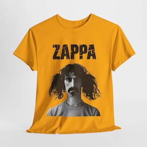 Puede incluir: Una camiseta amarilla con una imagen en blanco y negro de Frank Zappa y el texto "Zappa" en negro.