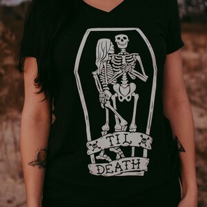 May include: Black V-neck t-shirt featuring a white graphic of two skeletons embracing inside a coffin. Below the coffin is a banner with the words "TIL DEATH" and skull and crossbones. The shirt is a dark color.