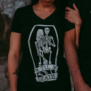 May include: Black t-shirts with a white graphic of skeletons in a coffin, with the text "Til Death". One shirt is a V-neck. The image shows a couple wearing the shirts.