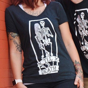 May include: Black t-shirts with a skeleton coffin graphic and the text "Till Death". The woman is wearing a v-neck shirt and black pants. The man is wearing a crew neck shirt and brown shorts.