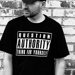 May include: A black short-sleeved t-shirt featuring a white rectangular graphic with the text "QUESTION AUTHORITY" and "THINK FOR YOURSELF" in bold capital letters, paired with a plain black baseball cap and dark denim jeans.