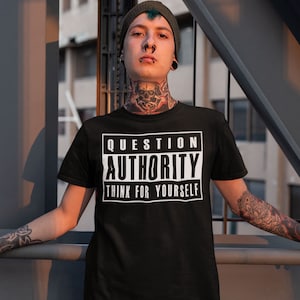May include: Black t-shirt with the text "QUESTION AUTHORITY THINK FOR YOURSELF" in a white, rectangular design. The shirt is worn by a person with tattoos and piercings, wearing a dark beanie.