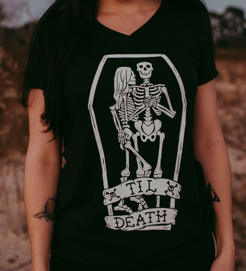 May include: Black V-neck t-shirt featuring a white graphic of two skeletons embracing inside a coffin. Below the coffin is a banner with the words "TIL DEATH" and skull and crossbones. The shirt is a dark color.