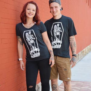 May include: Two people wearing matching dark gray t-shirts with a white skeleton design inside a coffin, with the text "Til Death". The person on the left is wearing black pants, and the person on the right is wearing brown shorts.