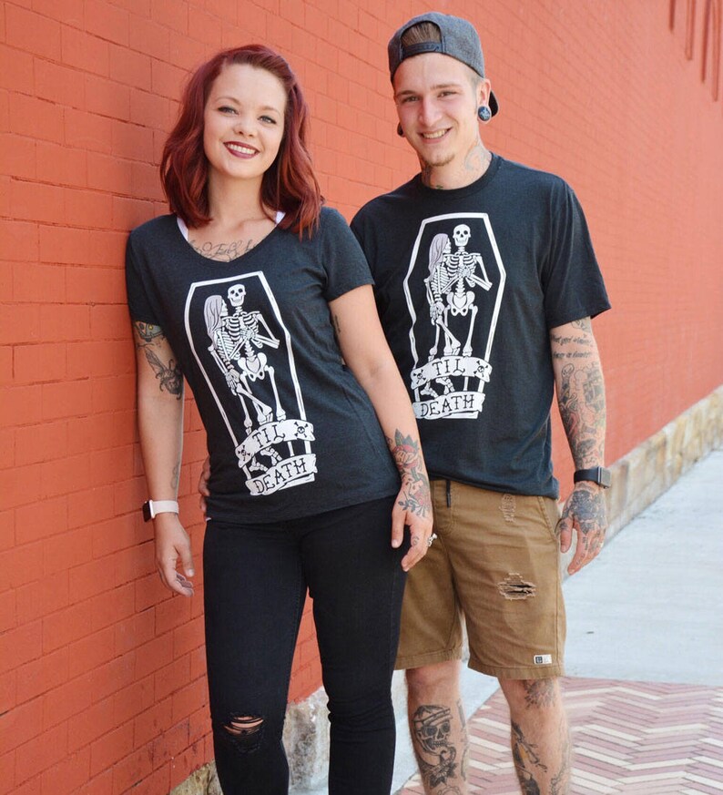 May include: Two people wearing matching dark gray t-shirts with a white skeleton design inside a coffin, with the text "Til Death". The person on the left is wearing black pants, and the person on the right is wearing brown shorts.