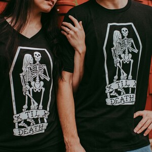 May include: Black t-shirts with a white graphic of skeletons embracing inside a coffin. The phrase "Til Death" is printed on a banner below the coffin. One shirt has a V-neck, the other has a crew neck.