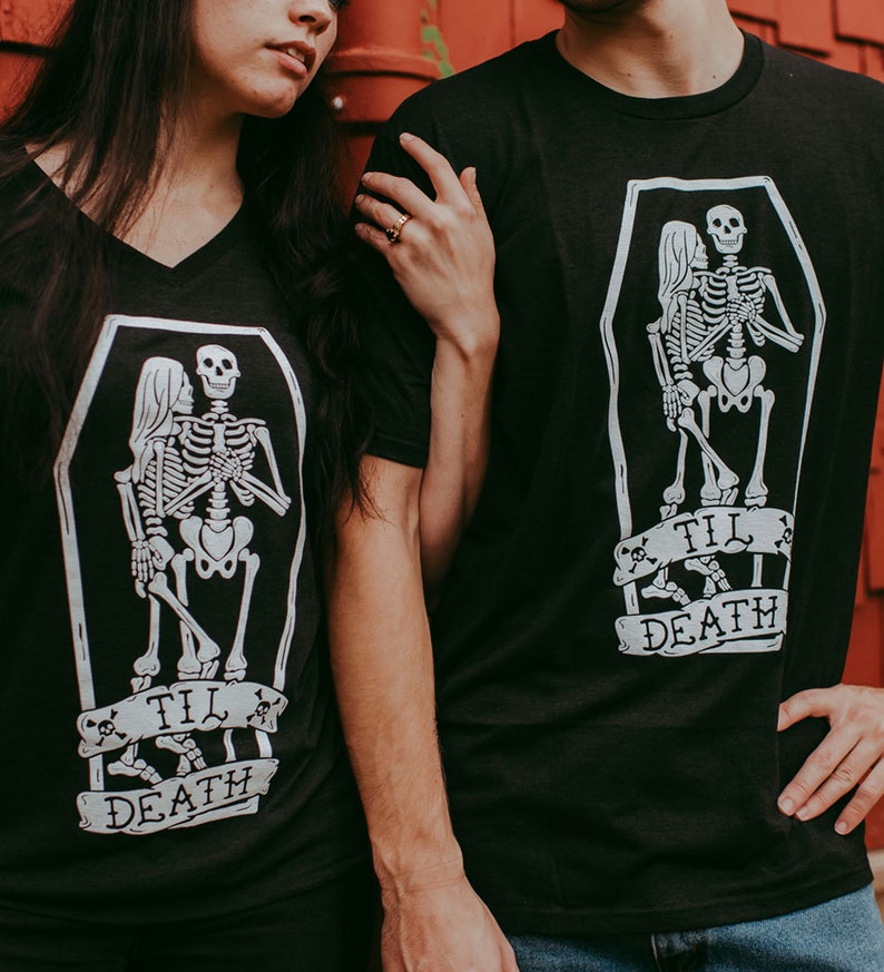 May include: Black t-shirts with a white graphic of skeletons embracing inside a coffin. The phrase "Til Death" is printed on a banner below the coffin. One shirt has a V-neck, the other has a crew neck.