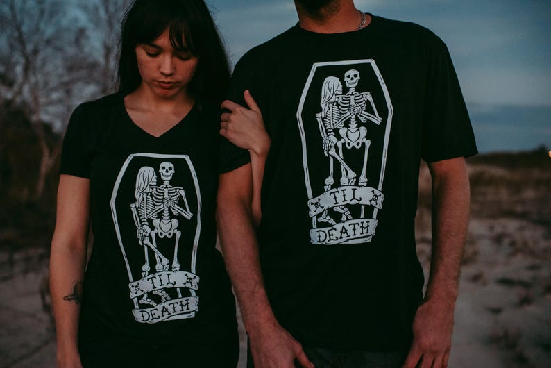 May include: Black t-shirts with a white graphic of skeletons in a coffin, with the text "Til Death". One shirt is a V-neck. The image shows a couple wearing the shirts.