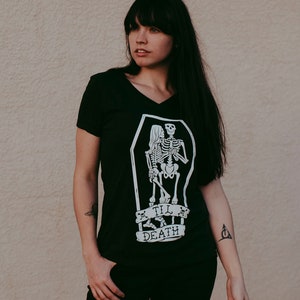 May include: Black V-neck t-shirt with a white graphic of two skeletons embracing inside a coffin. The text "Till Death" is below the coffin. The shirt is a dark color and has short sleeves.