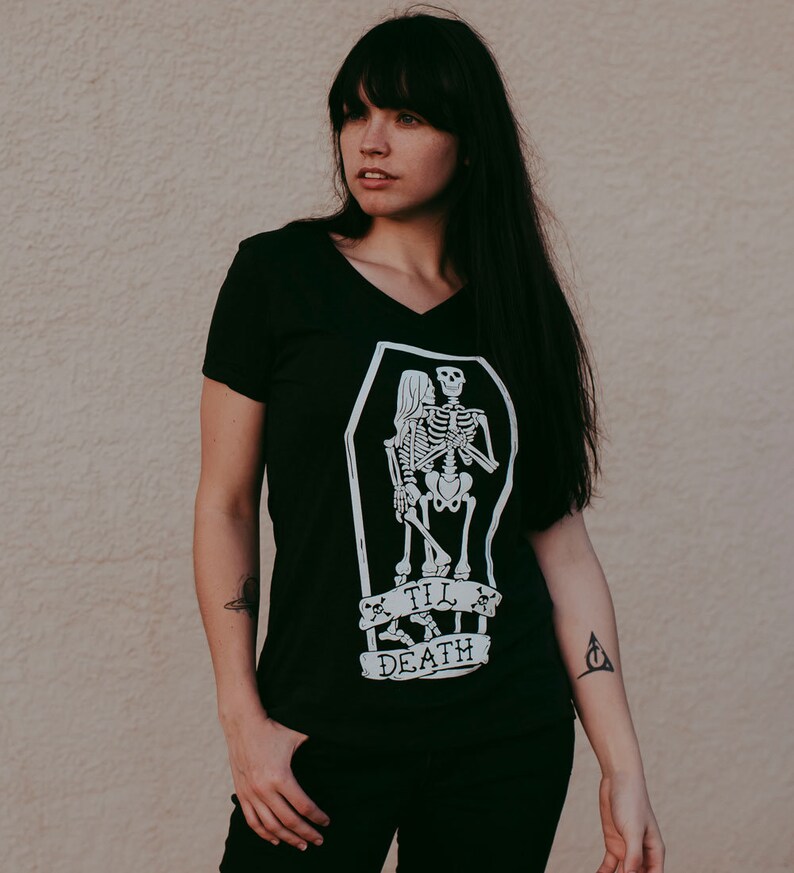 May include: Black V-neck t-shirt with a white graphic of two skeletons embracing inside a coffin. The text "Till Death" is below the coffin. The shirt is a dark color and has short sleeves.