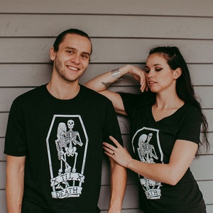 May include: Black t-shirts with a white graphic of skeletons in a coffin, with the text "Til Death". One shirt is crew neck, the other is a V-neck. The shirts are worn by two people standing in front of a gray wooden wall.