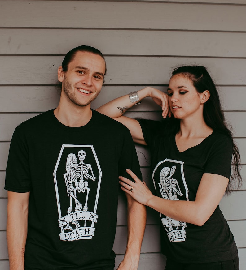 May include: Black t-shirts with a white graphic of skeletons in a coffin, with the text "Til Death". One shirt is crew neck, the other is a V-neck. The shirts are worn by two people standing in front of a gray wooden wall.