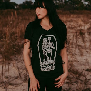 May include: Black V-neck t-shirt featuring a white graphic of two skeletons embracing inside a coffin. Below the coffin, a banner reads "Til Death". The shirt is a dark color and has short sleeves.