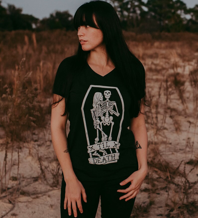 May include: Black V-neck t-shirt featuring a white graphic of two skeletons embracing inside a coffin. Below the coffin, a banner reads "Til Death". The shirt is a dark color and has short sleeves.
