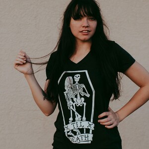 May include: Black V-neck t-shirt featuring a white graphic of two skeletons embracing in a coffin. The text "Til Death" is displayed below the coffin on a banner. The shirt is a dark color.