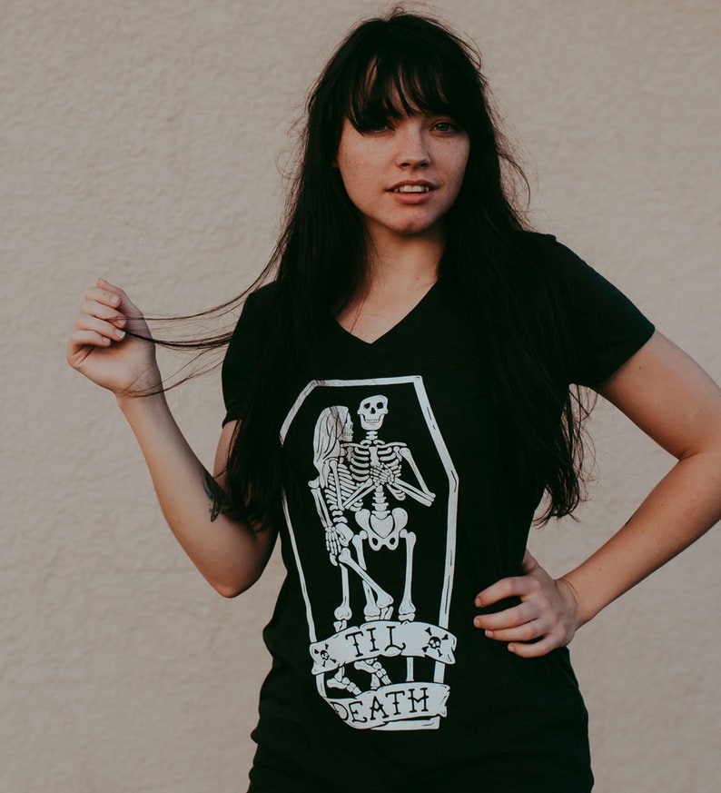 May include: Black V-neck t-shirt featuring a white graphic of two skeletons embracing in a coffin. The text "Til Death" is displayed below the coffin on a banner. The shirt is a dark color.