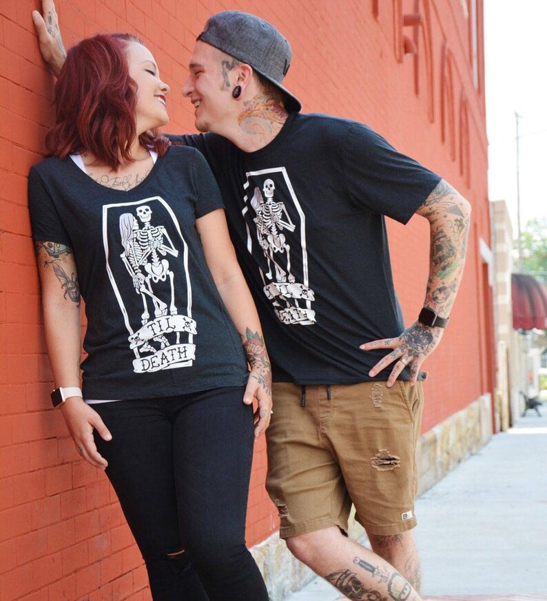 May include: Black t-shirts with a skeleton coffin graphic and the text "Till Death". The woman is wearing a v-neck shirt and black pants. The man is wearing a crew neck shirt and brown shorts.