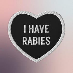 Puede incluir: Parche negro en forma de corazón con costuras blancas alrededor de los bordes. El parche dice "I HAVE RABIES" en texto blanco.