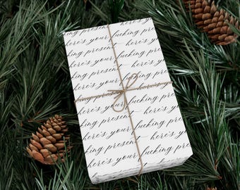 Könnte beinhalten: Ein verpacktes Geschenk mit weißem Papier mit dem Text "here's your fucking present" in einem sich wiederholenden Muster. Das Geschenk ist mit Bindfaden gebunden und liegt auf einem Bett aus grünen Kiefernadeln und Tannenzapfen.