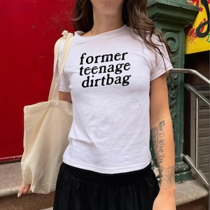 Könnte beinhalten: Weißes T-Shirt mit Rundhalsausschnitt und dem schwarzen Text "former teenage dirtbag". Das Shirt wird mit einem schwarzen Rock und einer beigen Tragetasche kombiniert. Der Hintergrund zeigt eine rote Tür und ein grün-gelbes Schild.