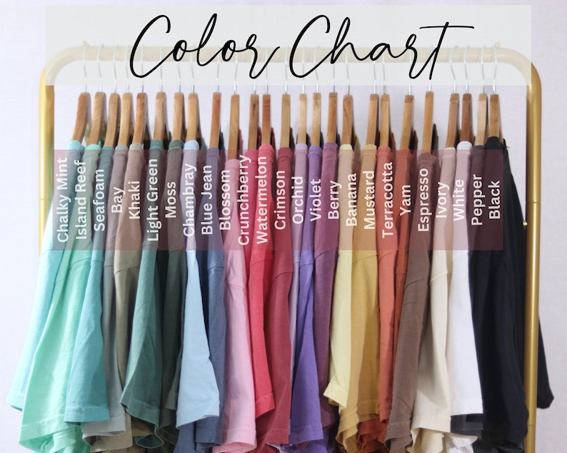 May include: A color chart displaying a row of t-shirts hanging on a gold rack. The shirts are labeled with text including Chalky Mint, Island Reef, Seafoam, Bay, Khaki, Light Green, Moss, Chambray, Blue Jean, Blossom, Crunchberry, Watermelon, Crimson, Orchid, Violet, Berry, Banana, Mustard, Terracotta, Yam, Espresso, Ivory, White, Pepper, and Black.