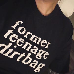 May include: A black short-sleeved t-shirt featuring the white text "former teenage dirtbag" printed in a distressed, serif font across the chest. The shirt has a classic crew neckline and a standard fit, showcasing a bold graphic design.