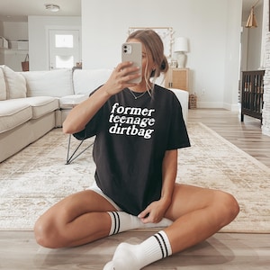 Könnte beinhalten: Ein schwarzes T-Shirt mit dem Aufdruck "former teenage dirtbag" in weiß auf der Vorderseite. Das Shirt wird von einer Person getragen, die auf einem Teppich sitzt.