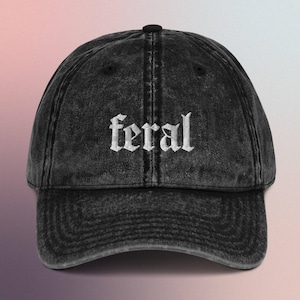 Puede incluir: Gorra de béisbol de denim negro desgastado con texto bordado en blanco que dice "feral" en una fuente gótica.