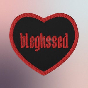 Può includere: Un cerotto a forma di cuore rosso con tessuto nero e la parola "bleghsed" ricamata in rosso.