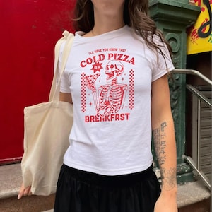 May include: White t-shirt with a red graphic of a skeleton holding a slice of pizza. The text on the shirt reads "I'll have you know that cold pizza is a real breakfast."