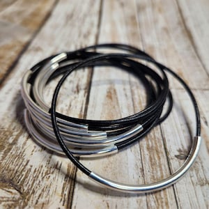 May include: A stack of black leather cord bracelets with silver-toned metal accents. The bracelets are coiled together, showcasing the contrast between the dark leather and the shiny metal. The background is a light brown wooden surface.