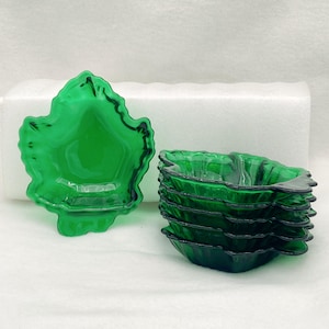 May include: A collection of green glass leaf-shaped bowls. One bowl is shown separately, with the others stacked. The bowls have a glossy finish and a dark green outline. These bowls are likely for serving snacks or small dishes.