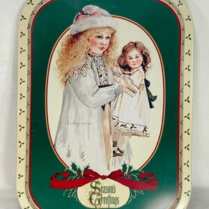 Könnte beinhalten: Ein vintage Serviertablett aus Metall mit grünem Hintergrund und weißem Rand. Das Tablett zeigt ein Gemälde von zwei Mädchen, eines hält eine Puppe, mit einem roten Band und Stechpalmenblättern am unteren Rand. Der Text "Season's Greetings" ist in einem goldenen Kreis gedruckt.
