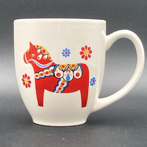 May include: A white ceramic mug with a black handle and the text "But First, Coffee" in a black, handwritten-style font. The mug is on a white surface.