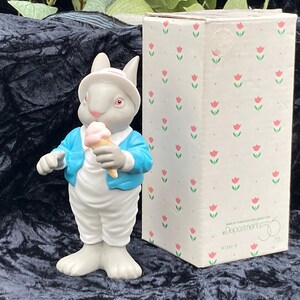 Puede incluir: Una figurita de conejo blanco que lleva una chaqueta azul y sostiene un cono de helado rosa y blanco. La figurita está de pie sobre una superficie negra frente a una caja blanca con un patrón floral rosa. La caja tiene el texto "Department 56" impreso en ella.