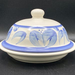 Op de afbeelding: Een witte keramische botervloot met een deksel. Het deksel en de basis zijn afgezet met een blauwe band. Het deksel is versierd met blauwe bloemmotieven. De schaal heeft een kleine, ronde knop bovenop.