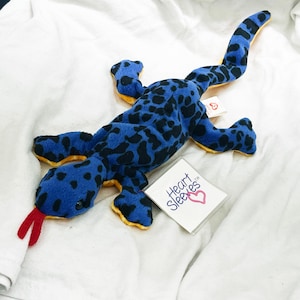 Puede incluir: Un juguete de lagarto de peluche con un cuerpo azul vibrante con manchas negras, una parte inferior amarilla y una lengua de fieltro roja. Una etiqueta blanca adjunta al costado muestra el texto Heart Sleeves con un icono de corazón rosa y una etiqueta roja Ty.