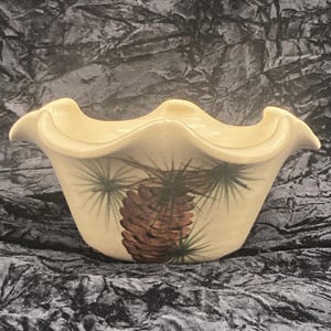May include: A white ceramic bowl with a wavy rim and a pine cone design. The bowl is decorated with a brown pine cone and green pine needles.