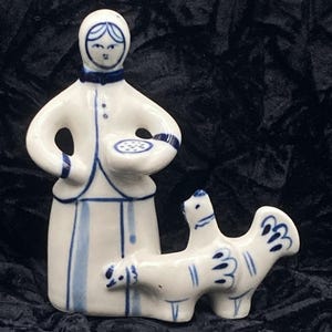 May include: A white ceramic figurine set featuring a woman holding a plate and a stylized animal. The figures are decorated with blue accents and lines. The woman wears a head covering and a long dress. The animal has a bird-like appearance.