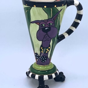 Puede incluir: Una taza de cerámica con forma de cono con una cara de perro púrpura en la parte delantera. La taza tiene un asa a rayas blancas y negras y se apoya en tres patas a rayas blancas y negras.