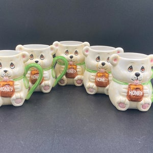 May include: Set of five ceramic mugs featuring a white bear design holding a honey pot. Each mug has a green handle and the word "Honey" is written on the honey pot.