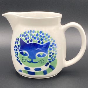 May include: White ceramic pitcher with a blue and green cat design. The cat's face is blue, surrounded by a green and blue dotted pattern. The pitcher has a spout and a curved handle.