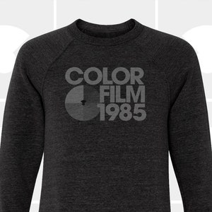 Puede incluir: Una sudadera gris oscuro con un estampado gráfico blanco que dice "COLOR FILM 1985" y una imagen estilizada de un rollo de película.