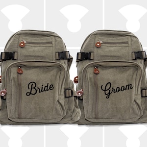 May include: Two canvas backpacks in a muted green color. One backpack is labeled "Bride" and the other "Groom" in black script. Each backpack has multiple pockets, zippers, and adjustable straps with black buckles and brown leather accents.