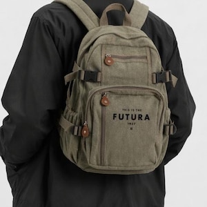 May include: Olive green canvas backpack with multiple pockets, adjustable straps, and a top handle. The backpack features the text "THIS IS THE FUTURA 1957" in black. The bag is worn by a person wearing a black jacket.