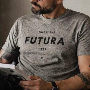 May include: A gray t-shirt with the text "THIS IS THE FUTURA 1927" in black. The person is wearing black-framed glasses and a black watch. The person is sitting in a black chair and reading a book.