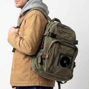 May include: An olive green canvas backpack with multiple pockets and adjustable straps. The backpack features a black graphic of a record and the text "Future rounds". The person in the image is wearing a brown jacket, blue jeans, and a black beanie.