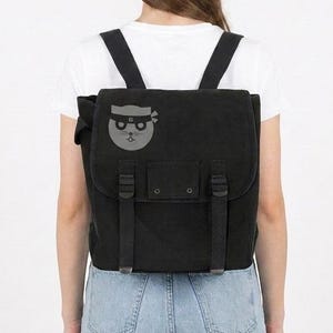 Puede incluir: Mochila negra con cierre de solapa, correas ajustables y bolsillo delantero. La mochila presenta un gráfico gris de un gato con una cinta de ninja. La persona de la imagen lleva una camiseta blanca y vaqueros azules.