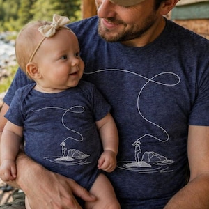 May include: A father and baby wearing matching navy blue shirts with a white fishing graphic. The father wears a tan baseball cap with a "SIMMS" patch. A fishing rod and reel are visible in the background.